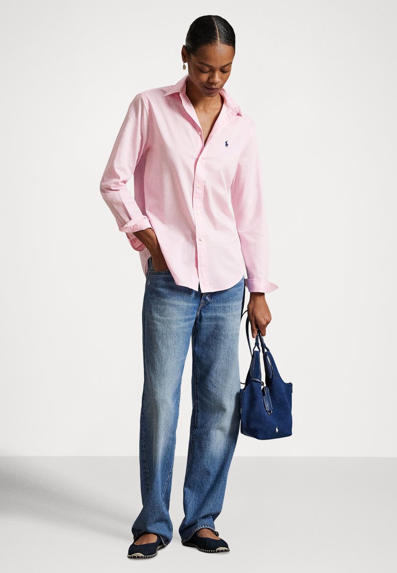 Donna con camicia rosa chiaro abbottonata, jeans blu larghi e ballerine nere, che tiene in mano una piccola borsa blu navy con un logo bianco.
