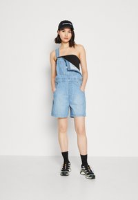Salopette in denim di colore azzurro chiaro con gambe corte, caratterizzata da una sola spallina, abbinata a una maglietta nera senza maniche e scarpe sportive nere.