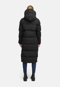 Black long puffer coat with a hood, featuring a quilted design, zip closure, and raised seams, worn over blue jeans and black boots.