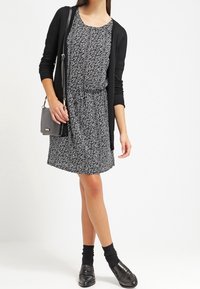 Schwarze Strickjacke über einem gemusterten schwarz-weißen Kleid mit Kordelzug in der Taille. Die Accessoires umfassen eine kleine Umhängetasche und schwarze Schuhe.