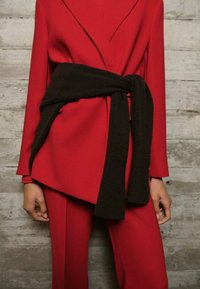 Red tailored suit with asymmetrical blazer and matching pants, paired with a black sweater tied around the waist against a concrete wall background.