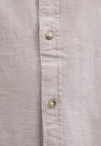 Camisa de lino gris claro con un acabado texturizado, que presenta una fila de botones perlados a lo largo del pliegue frontal. Tejido suave y transpirable.