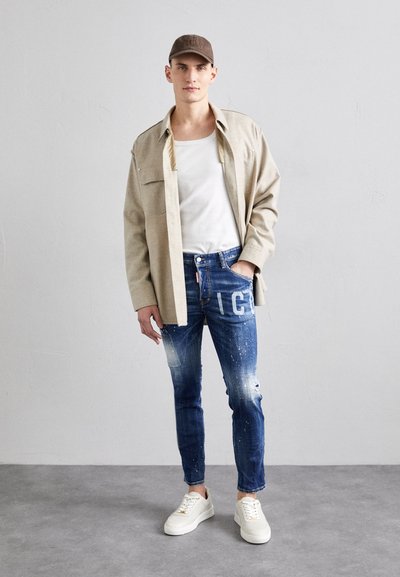 Beige übergroßes Hemd über einem weißen T-Shirt, kombiniert mit zerschlissenen blauen Jeans, die weiße Farbspritzer aufweisen. Weiße Sneaker runden das Outfit ab.