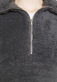 Sudadera gris de forro polar con un cuello alto, que tiene un cierre de cremallera plateada. Superficie suave y texturizada con un aspecto acogedor y grueso.