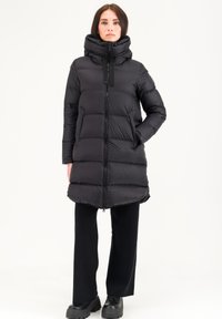 Doudoune noire avec un col montant et une fermeture éclair ; présente des panneaux matelassés horizontaux et des poches latérales ; portée sur un pantalon noir et des bottes.