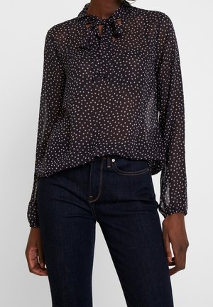 Kvinde iført en sort transparent bluse med små hvide prikker og en bundet sløjfe ved halsudskæringen, kombineret med mørkeblå jeans.