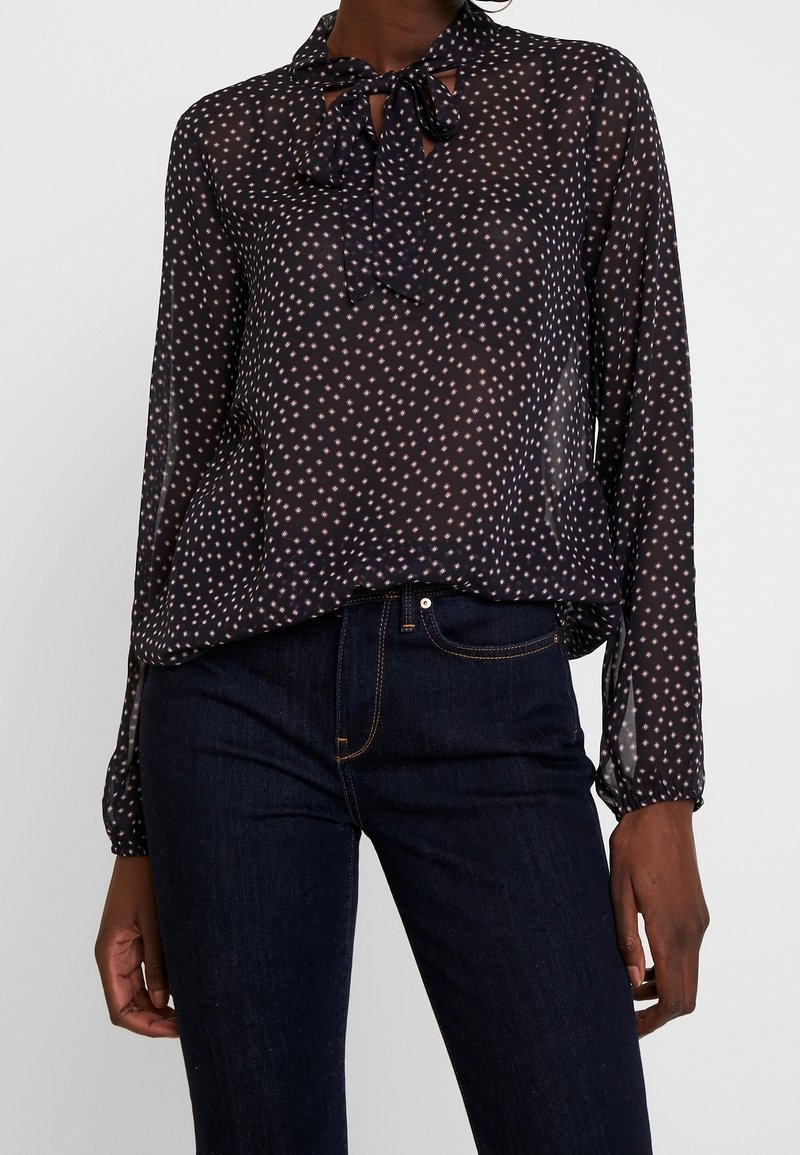 Femme portant un chemisier noir transparent avec de petits pois blancs et un nœud noué au niveau du col, associé à un jean bleu foncé.