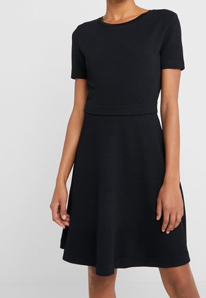 Femme portant une robe noire texturée à manches courtes, longueur genou, avec une taille cintrée et un col rond, devant un fond uni.