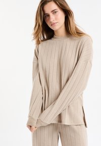 Beżowy prążkowany sweter dziany z wysokim kołnierzem i długimi rękawami, z bocznymi rozcięciami dla łatwości ruchu. Widoczna miękka faktura.