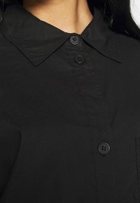 Gros plan d'une chemise noire à col avec trois boutons noirs fermés, montrant une partie du cou et des cheveux foncés d'une personne.