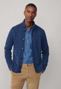 Blauwe kabelgebreide cardigan van textiel, met een geribde kraag en grote knopen, gecombineerd met een spijkerblouse en lichte corduroy broek.