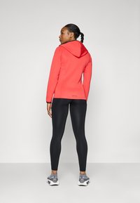 Rød hettejakke med svarte detaljer, tettsittende svarte leggings og grå treningssko. Glatt tekstur og strømlinjeformet design.
