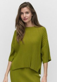 Neule, löysästi istuva villapaita kirkkaan vihreänä, jossa on pystysuoraa ribbikuvioa ja leveä kaula-aukko. Helma on hieman pidempi takana.