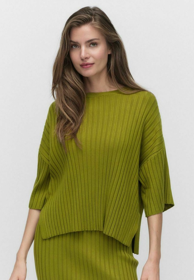 Neule, löysästi istuva villapaita kirkkaan vihreänä, jossa on pystysuoraa ribbikuvioa ja leveä kaula-aukko. Helma on hieman pidempi takana.