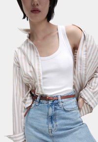 Weißes, geripptes Tanktop unter einem hellen, gestreiften Hemd mit hochgekrempelten Ärmeln. Hellblaue, hoch geschnittene Denim-Shorts und ein brauner Ledergürtel.