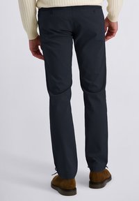 Marineblauwe broek met een gladde textuur, voorzien van twee achterzakken en een elastische zoom. Gecombineerd met lichtbruine suède schoenen.