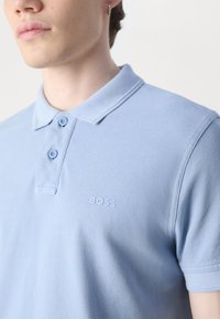 Šviesiai mėlyna polo marškinėlių, pagamintų iš tekstūruoto audinio, klasikinio apykaklės, su dviem mygtukais ir subtiliu toniniu "BOSS" logotipu ant krūtinės.