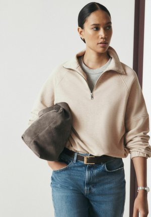 Kvinde med mørkt hår iført en beige kvart-lynlåssweatshirt og blå jeans, bærende en stor grå blød taske under den ene arm, kigger til siden.