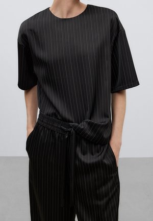 Persona que lleva una camiseta negra de manga corta con rayas finas y pantalones a juego con cintura ajustable, manos en los bolsillos, sobre un fondo liso.