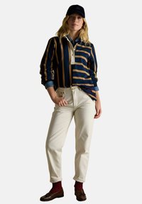 Woman wearing a navy and tan striped long-sleeve shirt, cream high-waist pants, burgundy socks, brown loafers, and a navy cap.