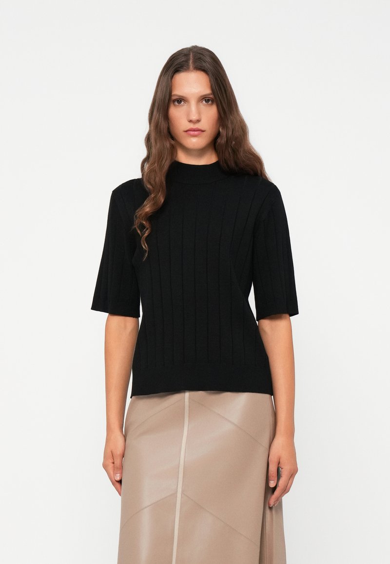 Zwarte geribbelde gebreide trui met korte mouwen en een hoge hals, gecombineerd met een beige geplooide rok. Zachte textuur en slim fit ontwerp.