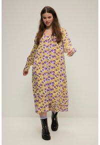 Vestido largo de botones con un fondo amarillo y patrones florales en púrpura y blanco. La modelo lleva puestas botas de tobillo negras con cremallera.
