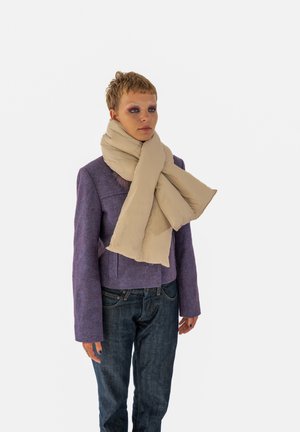 Beige pufi sál a nyakon, texturált anyag; egy lila dzsekivel, kék farmernadrággal és semleges háttérrel párosítva.