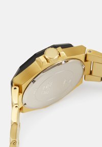 Gullfarget metallklokke med svart bezel, fremtredende krone og polert sølvbunn. Har en metallrem med lenkedesign.