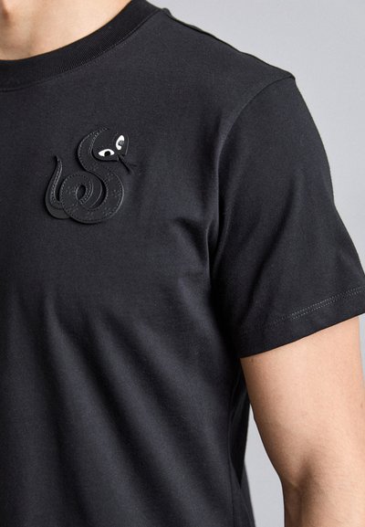 Zwarte katoenen t-shirt met een getextureerd, geborduurd slangontwerp op de bovenste linkerborst met witte ogen. Korte mouwen en een gladde afwerking.