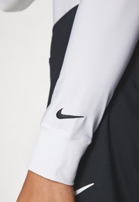 Dlouhé rukávy sportovní tričko z bílé látky, s ribbovanými manžetami a černým logem Nike na rukávu. Má elegantní, slim fit design.