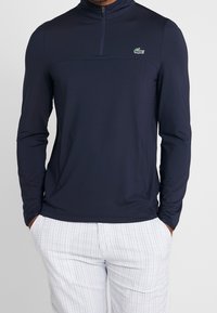 Marinblå långärmad tröja med hög krage och halvdragkedja, med ett strukturerat tyg och en liten grön logotyp på bröstet.