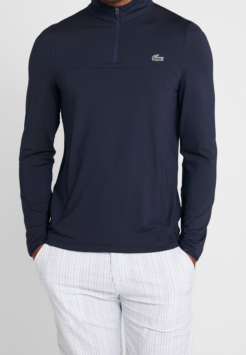 Marinblå långärmad tröja med hög krage och halvdragkedja, med ett strukturerat tyg och en liten grön logotyp på bröstet.