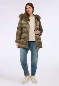 Oliwkowa kurtka puchowa z futrzanym kapturem, pikowaną fakturą, przednim zamkiem błyskawicznym i bocznymi kieszeniami, zestawiona z beżową bluzą i niebieskimi dżinsami.