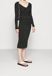 Robe midi noire à côtes avec manches longues et décolleté en V profond, associée à des sandales noires et blanc cassé. Design lisse et ajusté.