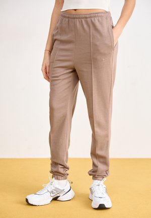Pantalon jogger marron coupe décontractée avec coutures à l'avant, poignets élastiques et poches latérales, porté avec des baskets blanches Nike et un top blanc court.