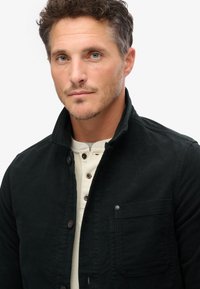 Zwarte corduroy jas met een hoge kraag, voorkeurszak en bruine knopen over een lichtbeige henley shirt. De textuur lijkt zacht en duurzaam.