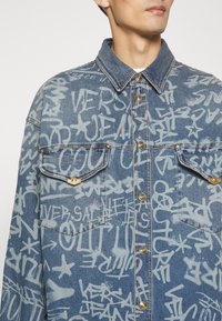 Traperica s lagano plavom podlogom i bijelim natpisima u stilu grafita. Ima šiljastu kragnu, dva džepa na prsima i zlatne dugmadi.