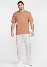 Vīrietis stāv brūnā Nike T-kreklā, baltās biksēs un baltās kedās pret vienkāršu baltu fona.