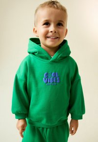 Mosolygó fiatal gyermek rövid szőke hajjal, zöld kapucnis pulóvert és nadrágot visel, a kapucnis pulóver felirata: "COOL VIBES HAVE FUN!"