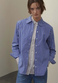 Blau-weiß gestreiftes Hemd mit Knöpfen und großem Kragen, darüber ein weißes Hemd mit dünnen schwarzen Streifen. Legerer Schnitt und lange Ärmel.