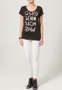 Woman wearing black T-shirt with white text, white rolled-up pants, black open-toe ankle boots, and a black beaded bracelet on left wrist.