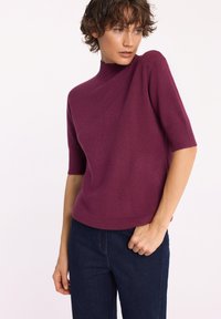 Krótka bluzka z krótkim rękawem, w kolorze głębokiego fioletu, z wysokim kołnierzem, z prążkowanymi mankietami i dołem, w zestawie z ciemnymi dżinsami. Gładka tekstura, codzienny krój.