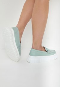 Světlé mátové zelené slip-on boty s texturovaným svrškem, které mají silnou bílou gumovou podrážku a dekorativní pásek přes přední část.
