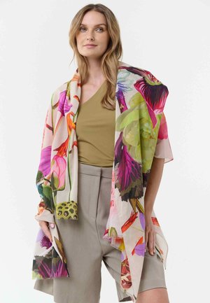 Vrouw met lang blond haar, gekleed in een olijfgroen topje en grijze korte broek, omhuld met een grote kleurrijke sjaal met bloemenpatroon.