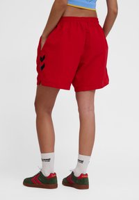 Röda shorts med elastisk midja, sidofickor och svarta chevron-accenter på vänster sida. Tillverkade av lätt material.