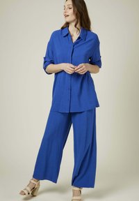 Chemise en lin bleu avec un col et des boutons, associée à un pantalon bleu ample. Texture lisse, coupe décontractée et design à jambes larges. Des sandales complètent la tenue.
