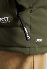 Olivově zelená kapsa na bundě s černým táhlem zipu označeným "JACK1T" a bílým vyšíváním "DCC" a částečným textem "JACK1T".