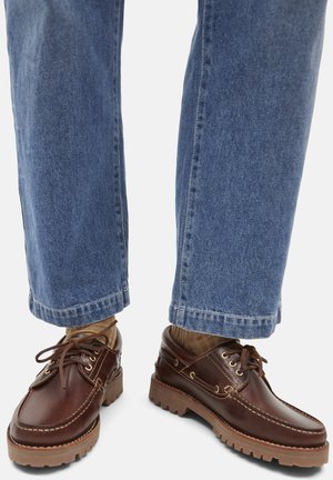 Person wearing blue jeans, brown socks, and dark brown leather lace-up shoes with thick rubber soles on a white background.