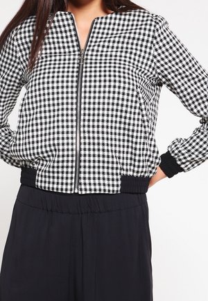 Vrouw draagt een zwart-witte ruitjesjas met ritssluiting, geribbelde boorden en zoom, gecombineerd met hoogwater zwarte broek.