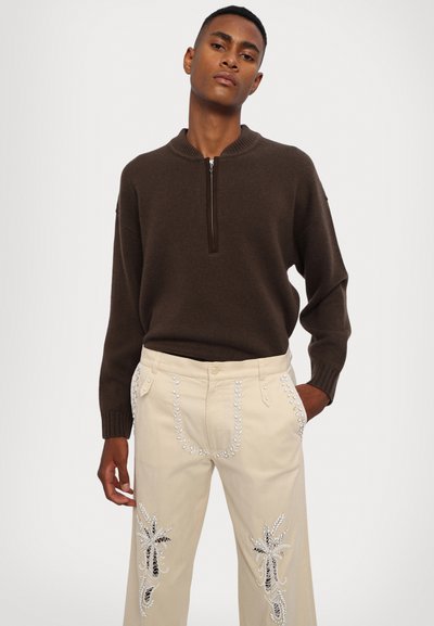 Maglione marrone con collo a zip in maglia morbida, abbinato a pantaloni beige chiaro con intricati ricami bianchi e hardware decorativo.